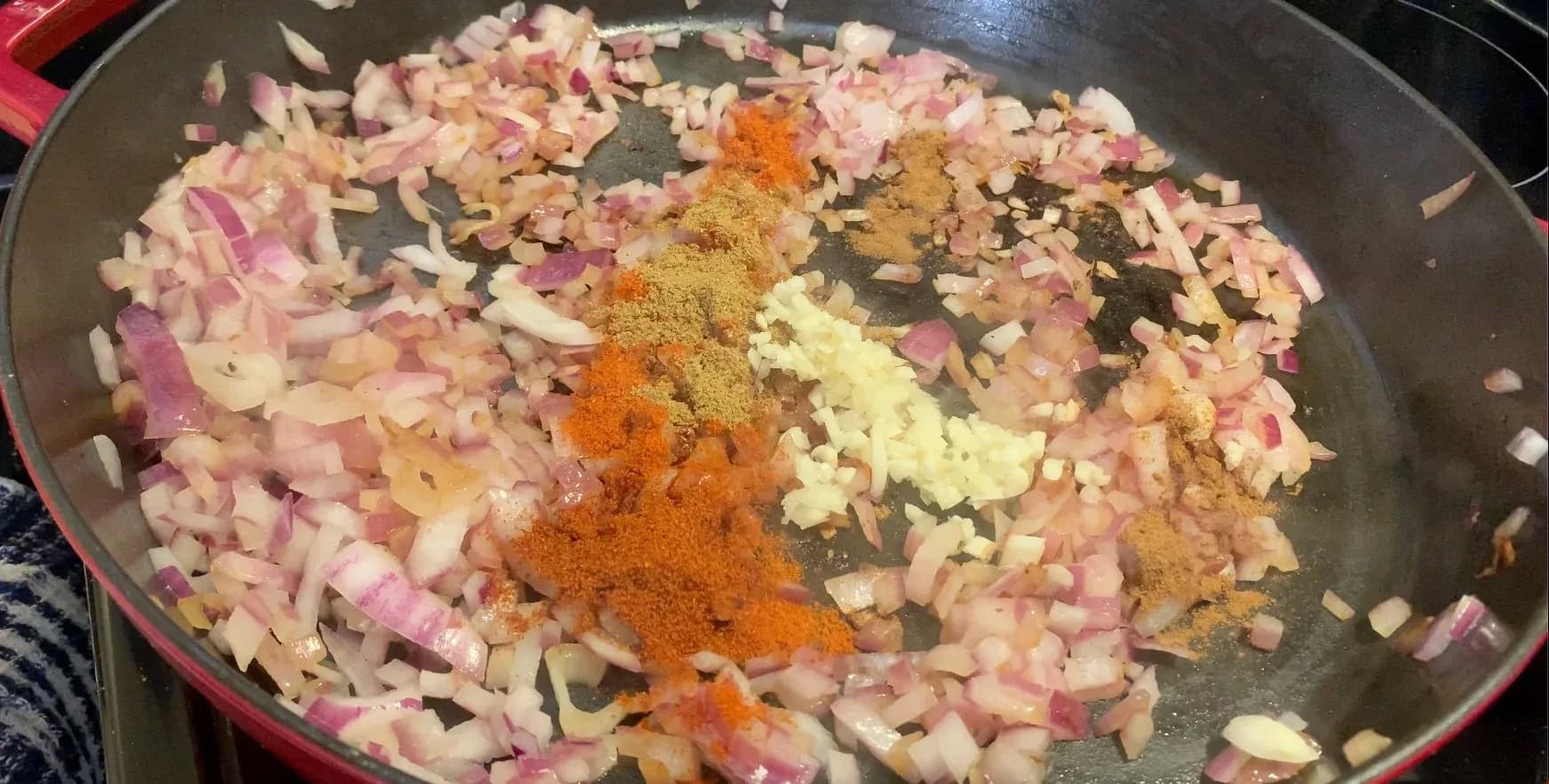 Diced onions and spices including paprika, cayenne, cinnamon, cumin, garlic, and ginger being sautéed in a large pan.