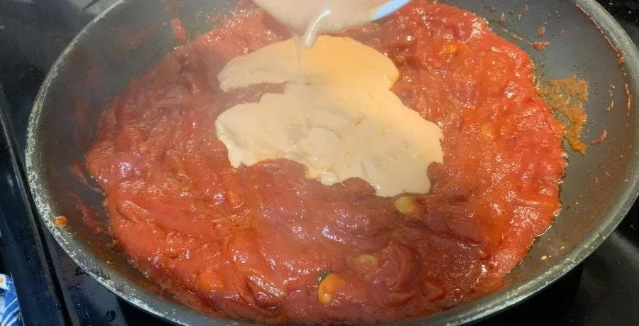 Red tomato sauce simmering in a pan with a dollop of yogurt being added to temper it for Shahi Paneer.
