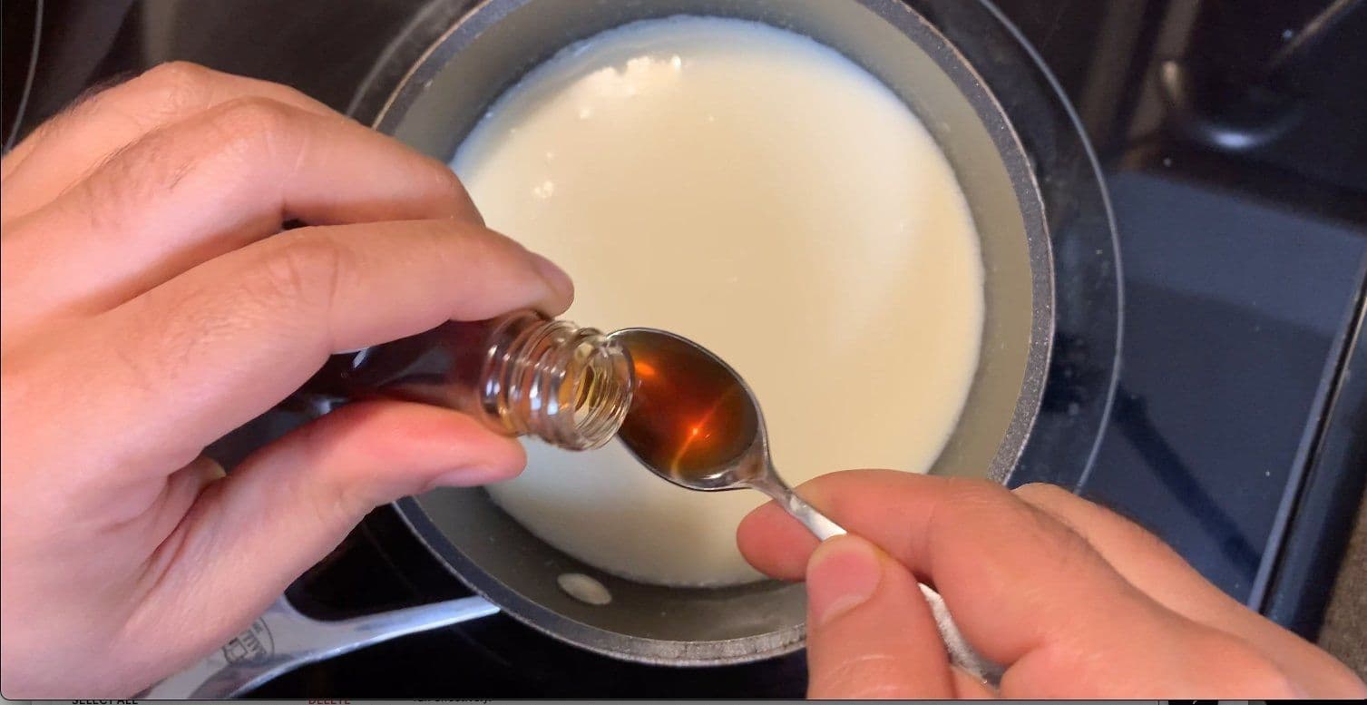 Pouring vanilla extract from a small bottle into a pot of melted white chocolate and milk.
