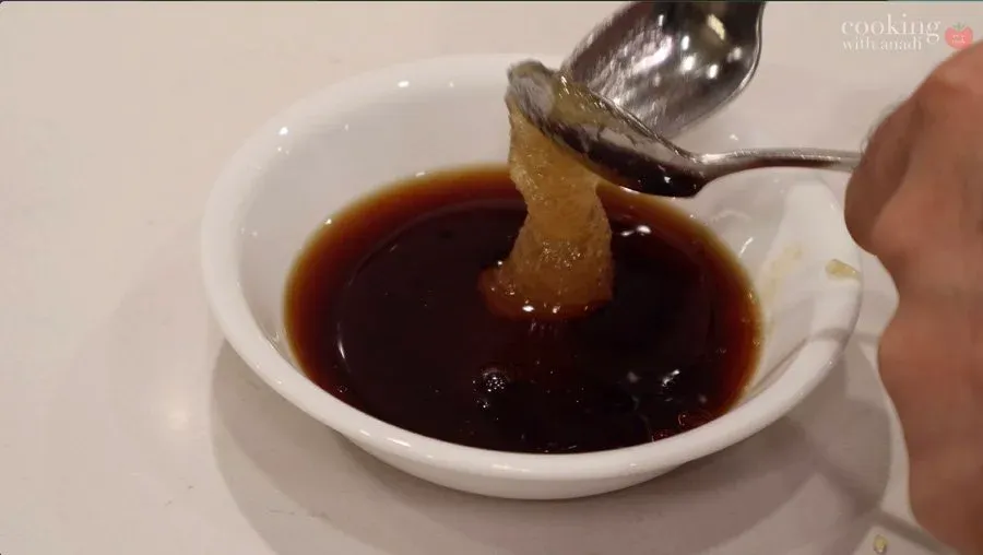 Spoon lifting a piece of ginger from a dark brown stir fry sauce in a white bowl.