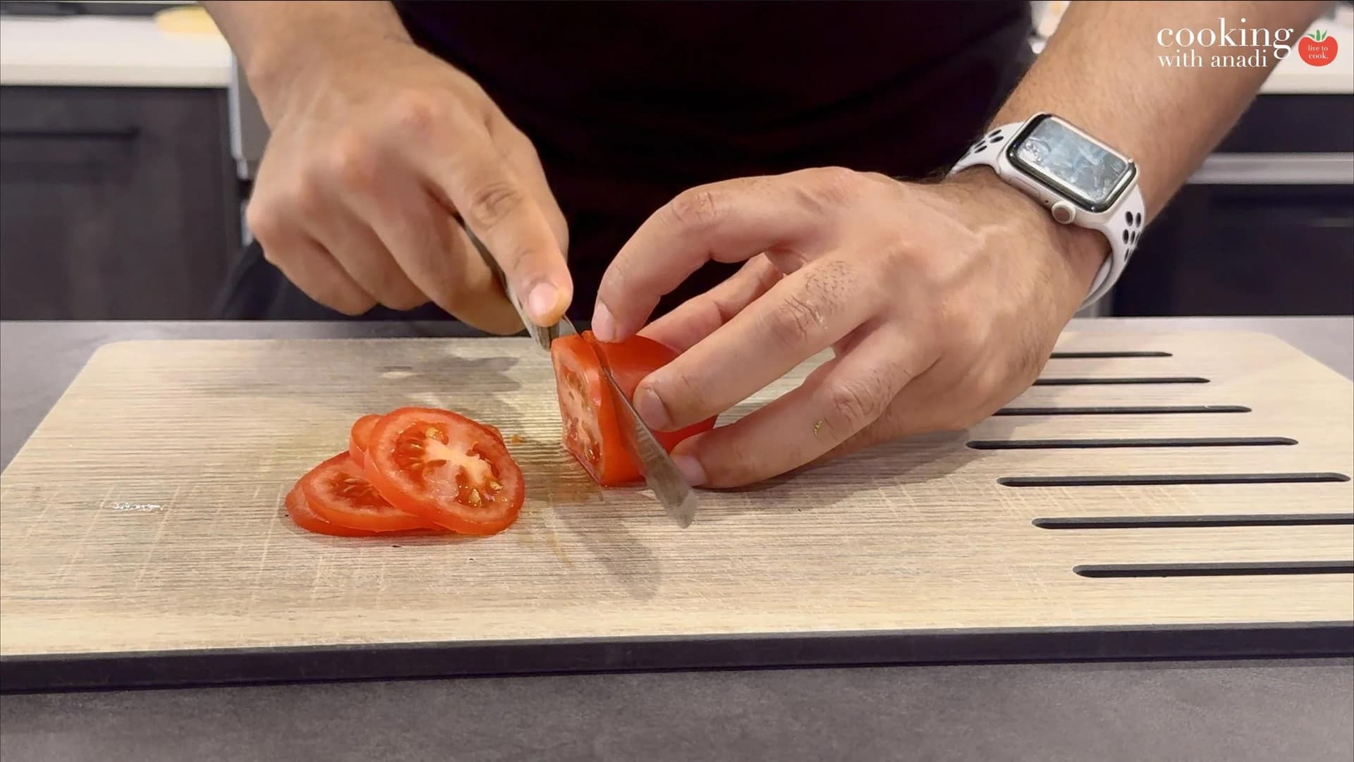 Hands slicing a Roma tomato into thin rounds on a cutting board, with several slices already cut nearby.