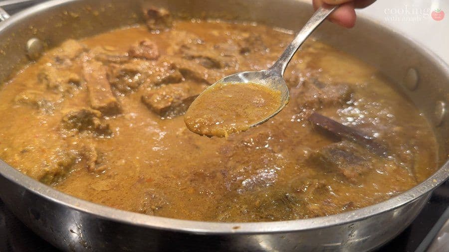Spoon lifting a serving of rich, brown Balinese beef rendang from a simmering pot of tender meat in coconut curry sauce.