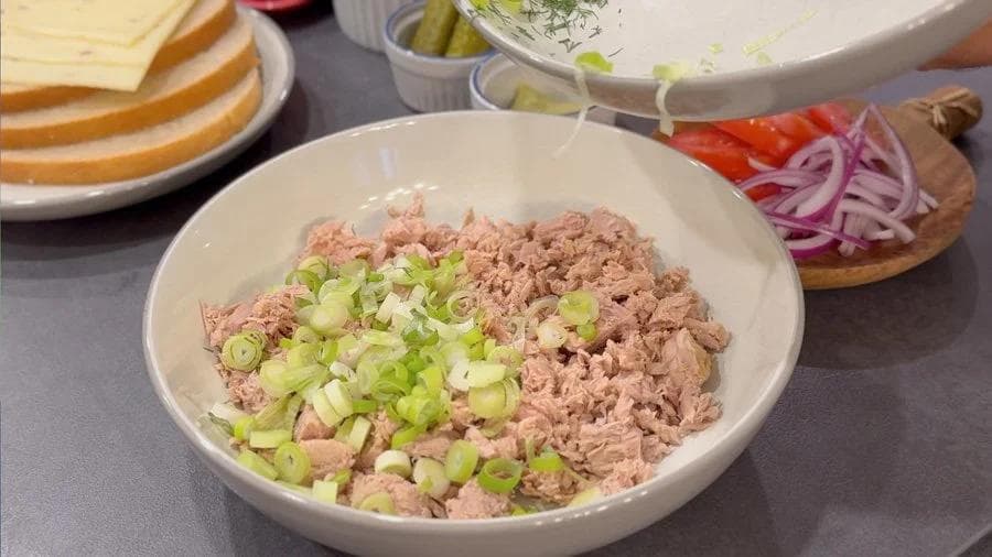 Drained canned tuna mixed with chopped green onions in a white bowl, with ingredients and bread visible nearby.