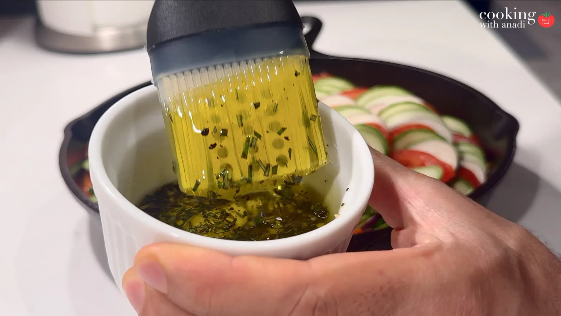 Silicone basting brush being dipped into a white ramekin of herb-infused olive oil with visible spices and green herbs.