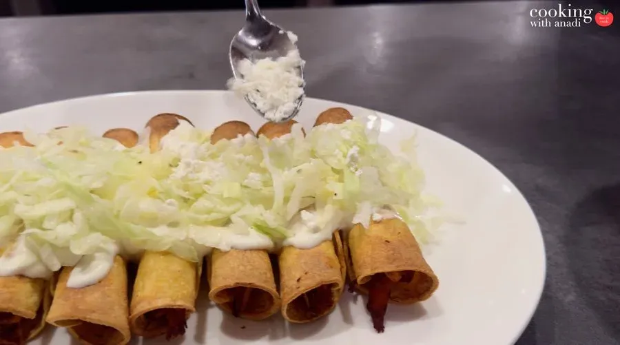 Crispy golden taquitos on a white plate, topped with sour cream, shredded lettuce, and cotija cheese being spooned on top.