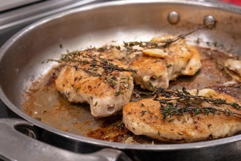 Pan-fried chicken breasts with fresh thyme sprigs in a stainless steel skillet with browned pan sauce.