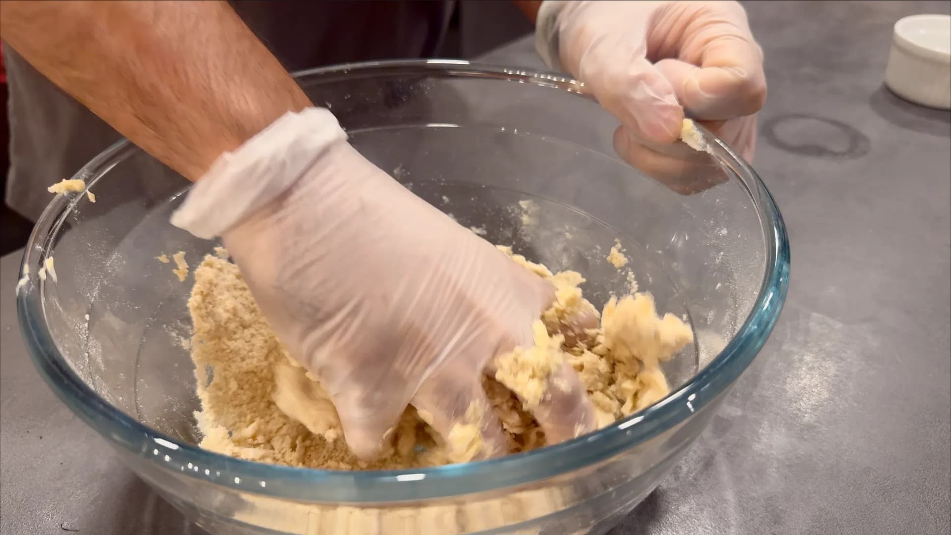 Hands in disposable gloves mixing dough ingredients in a glass bowl for Nigerian meat pie filling.