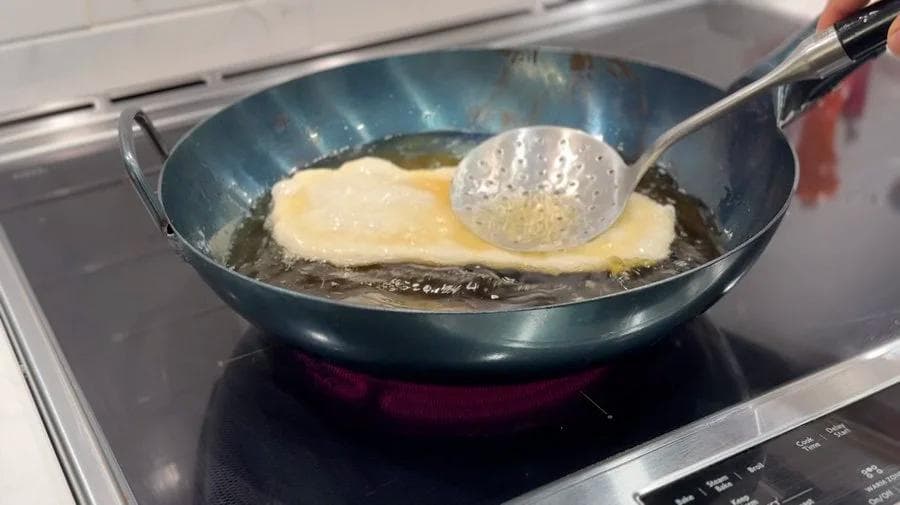 Deep-frying a stretched dough pastry in hot oil using a slotted spoon to press it down in a wok.
