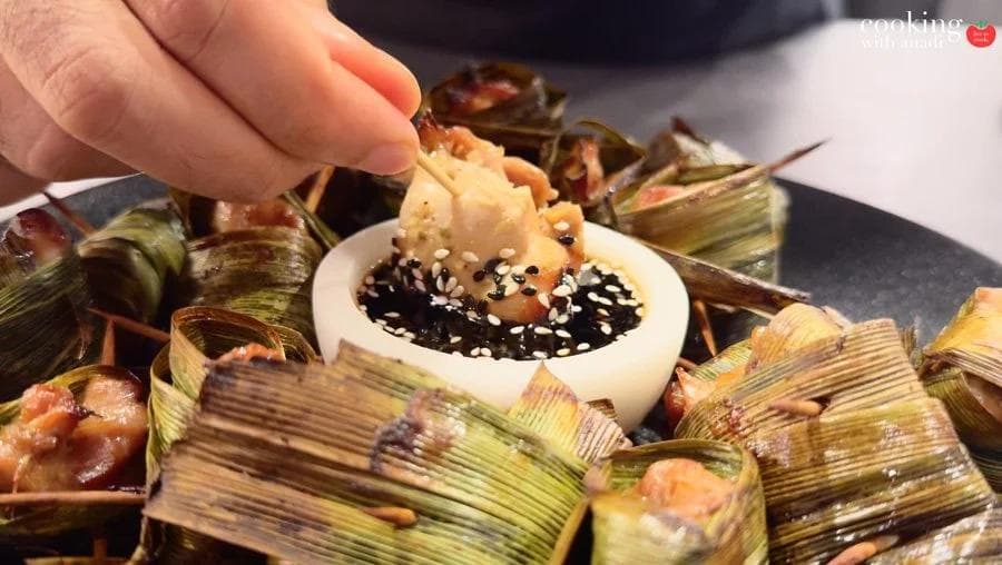 Pandan-wrapped chicken pieces arranged on a dark plate, one being dipped into a dark soy dipping sauce with sesame seeds.