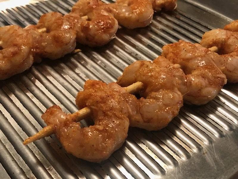 Marinated shrimp skewers cooking on a ridged grill pan, showing a reddish tandoori spice coating before flipping.