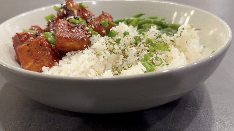 Crispy Korean-style tofu bites over sushi rice, garnished with green onions and sesame seeds in a white bowl.