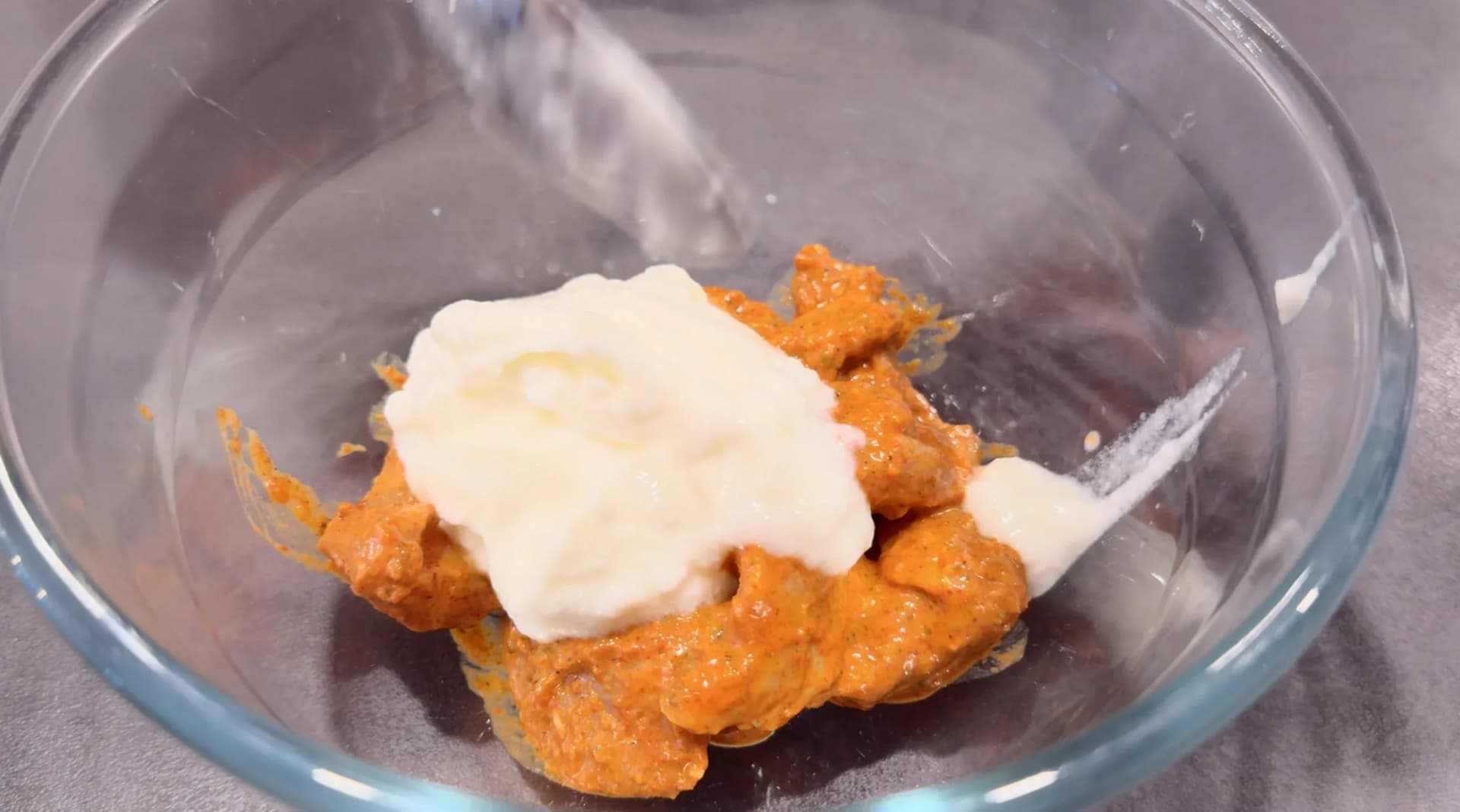 Yogurt being added to spiced orange chicken tikka marinade in a clear glass mixing bowl.