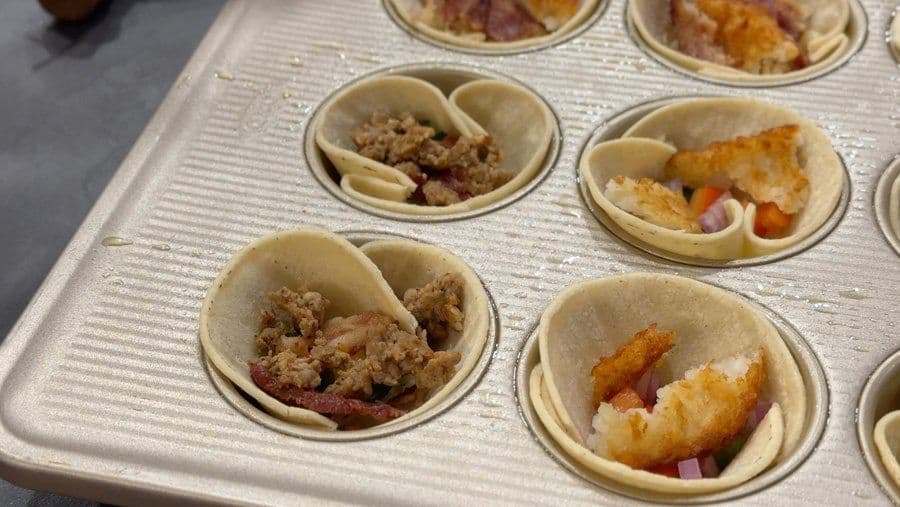 Unbaked savory egg cups in a muffin tin filled with meat, cheese, and hash browns before baking.