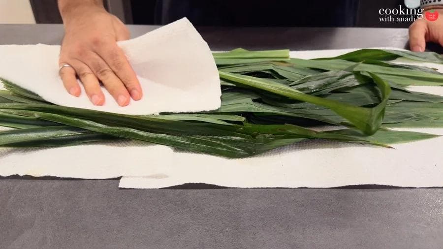 Hands patting fresh pandan leaves dry with paper towels on a flat surface.