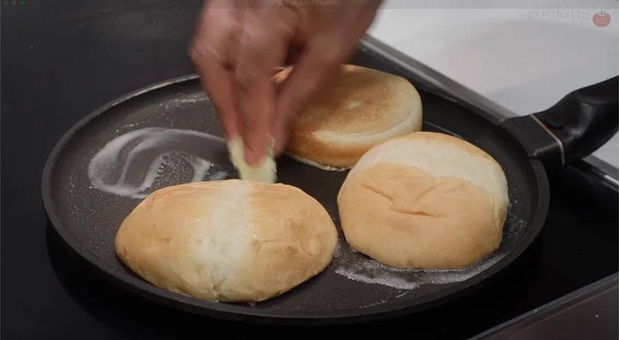Toasting burger buns in a black skillet on a stovetop, with a hand holding one bun.