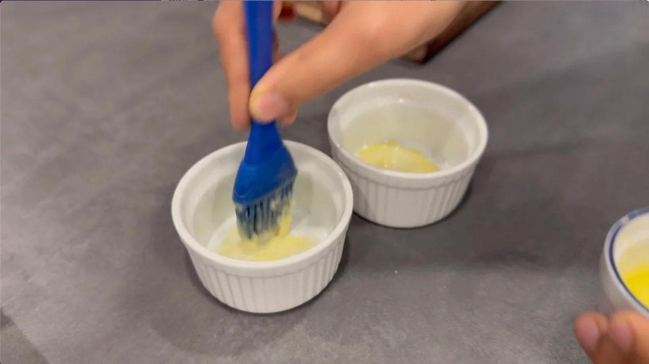 Brushing melted butter into white ramekins with a blue pastry brush for cheese soufflé preparation.