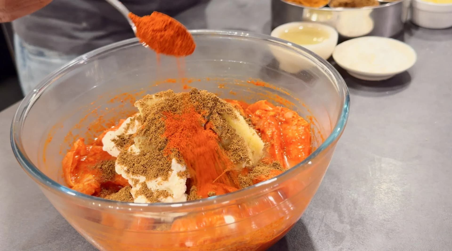 Red spice being spooned into a glass bowl of chicken coated in orange marinade, yogurt, and ground spices.