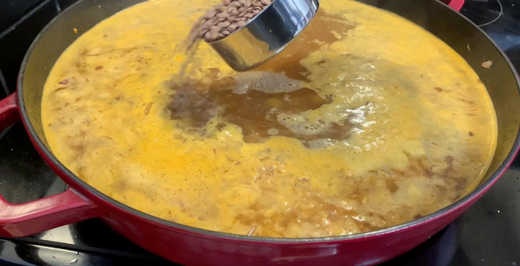Golden broth being stirred into a red pot of Moroccan chicken stew with visible chickpeas and lentils.