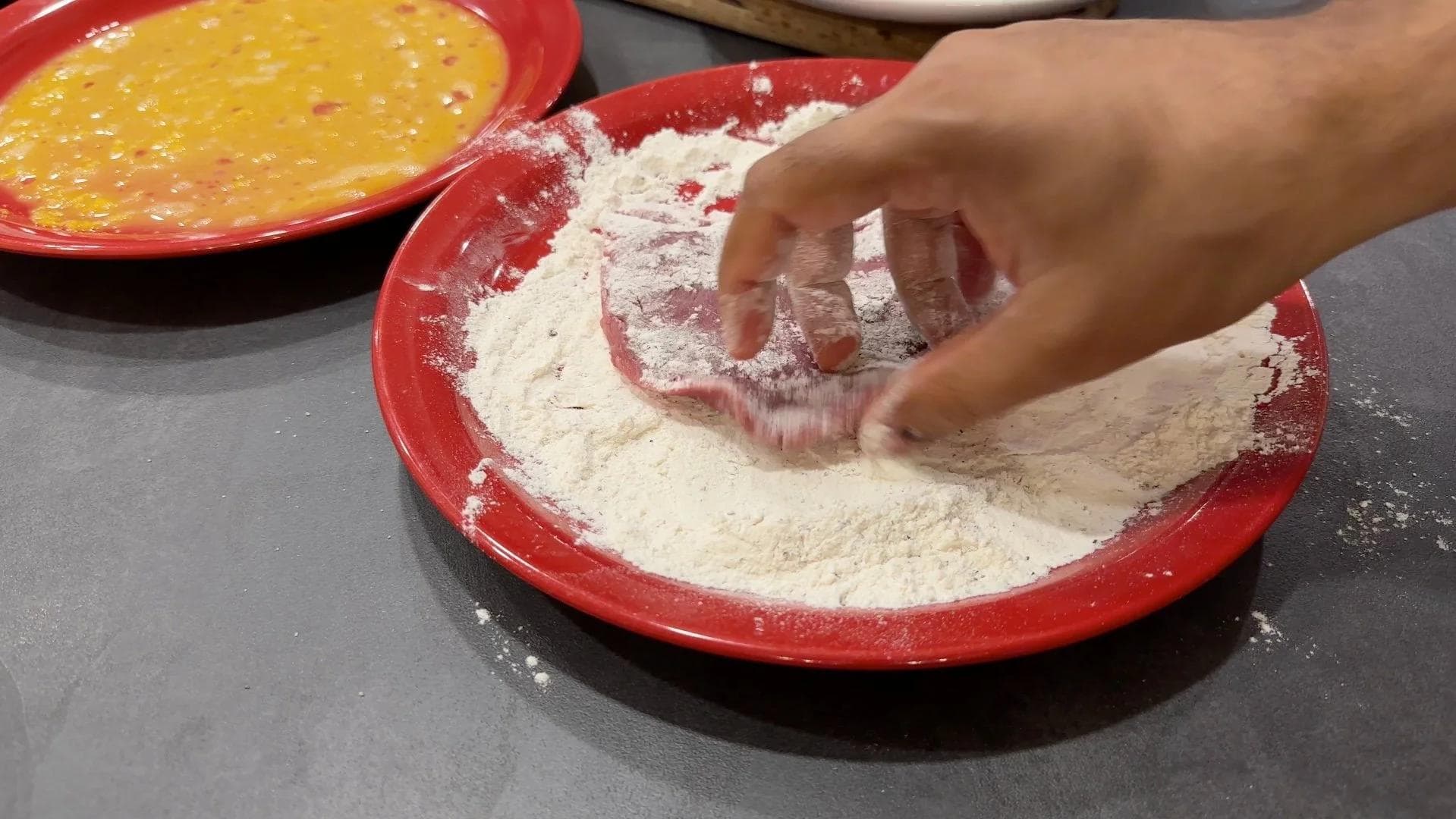 Hand dredging veal cutlet in flour on red plate during wiener schnitzel preparation.