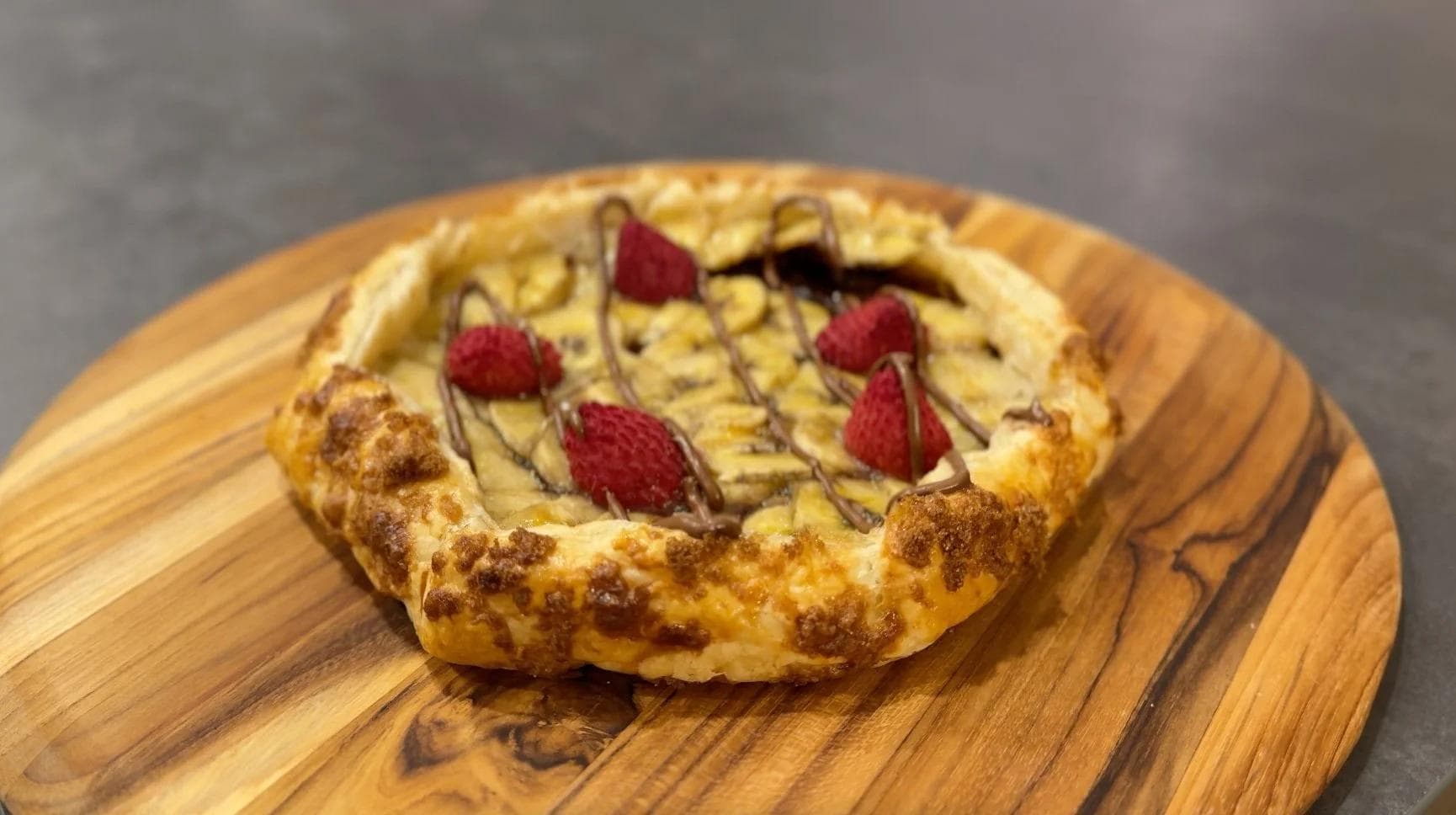 Baked banana Nutella galette topped with fresh raspberries and chocolate drizzle on wooden board.