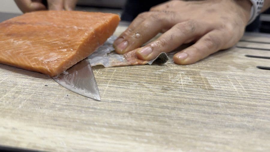 Removing skin from salmon fillet with a long knife on a wooden cutting board.