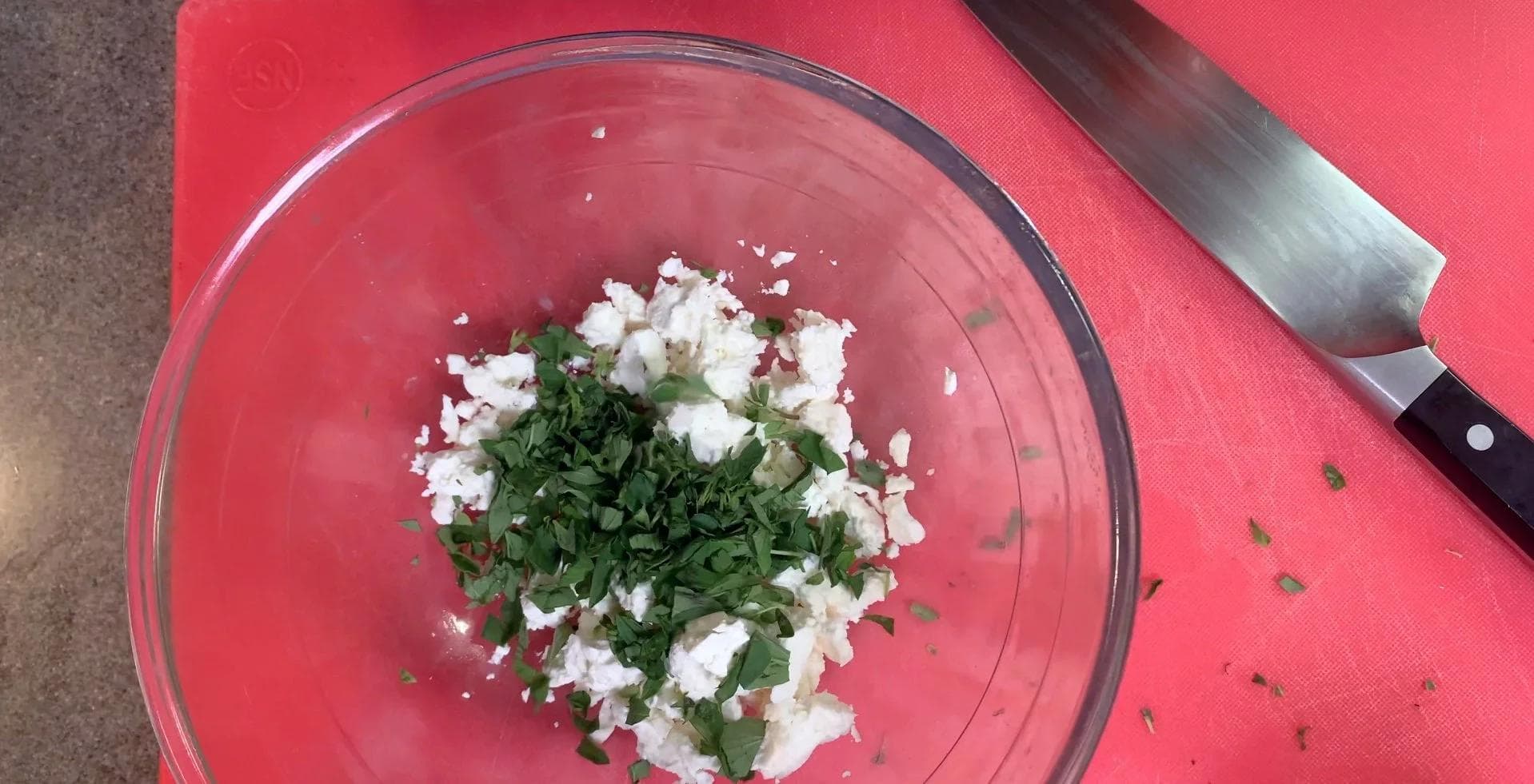 Crumbled feta cheese mixed with fresh oregano leaves in a glass bowl on a red surface with a knife nearby.
