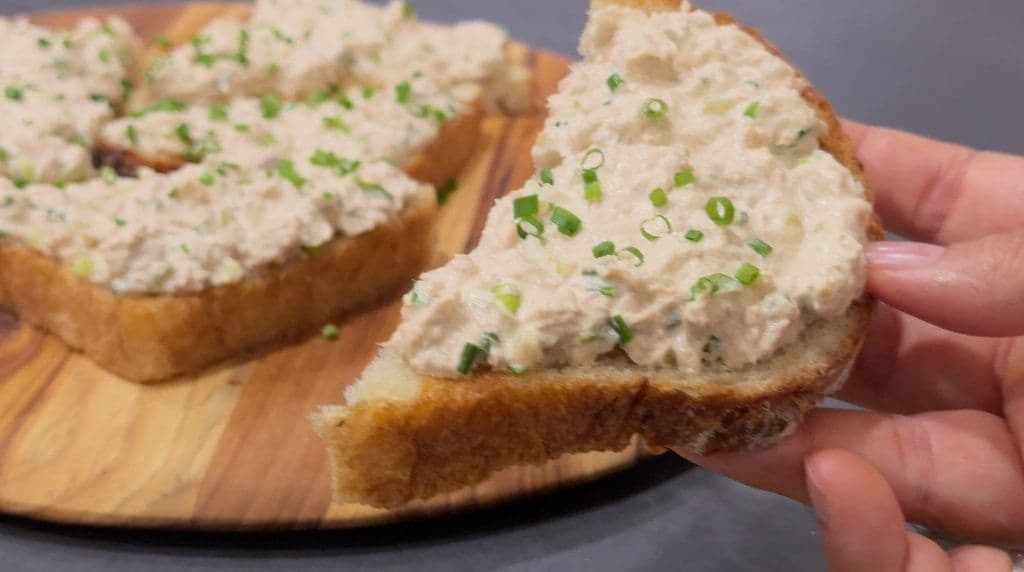 Hand holding a slice of toasted bread topped with creamy tuna salad and garnished with fresh green onions.