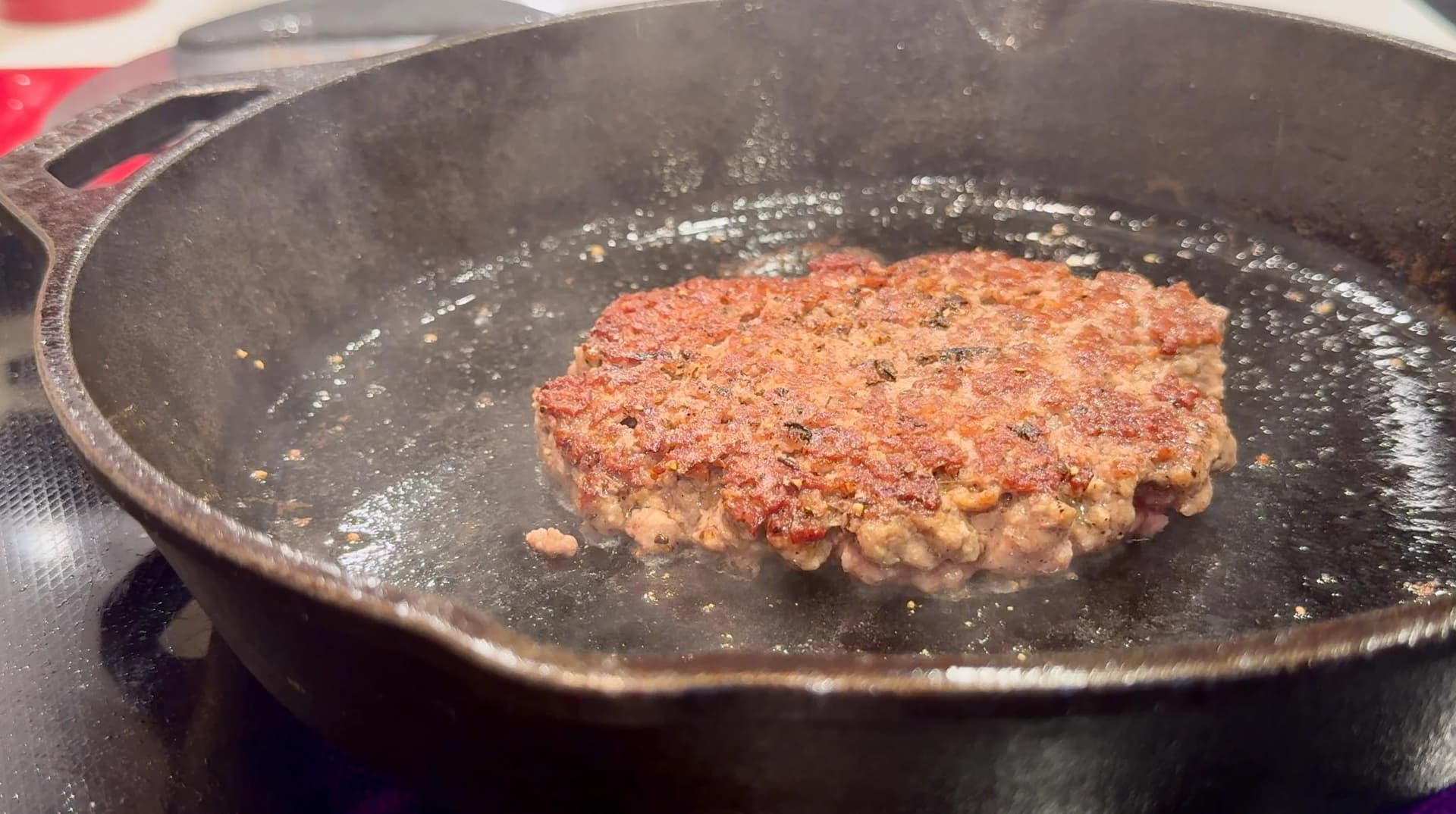 A seasoned beef patty searing in a cast-iron skillet, its top edge deep brown and craggy with a freshly seared crust