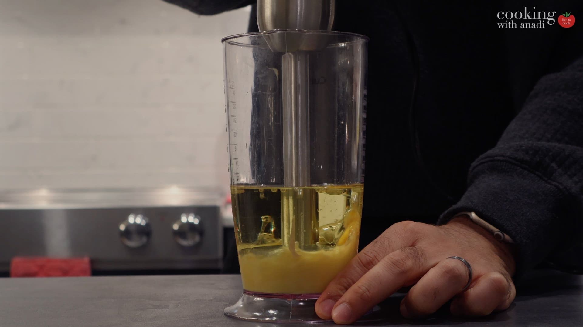 Immersion blender positioned at bottom of glass container with oil layered on top of mayonnaise ingredients.