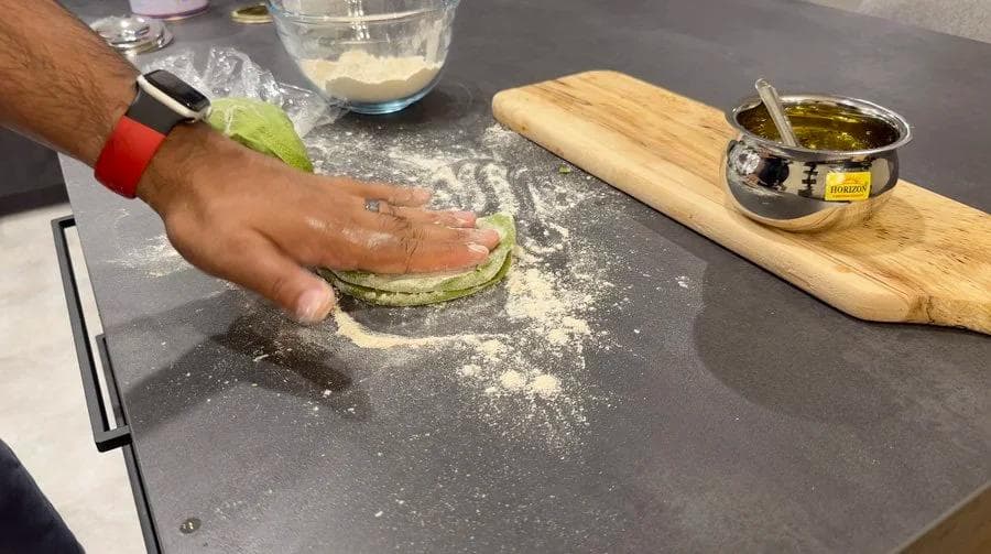 Hand folding a green spinach paratha dough in half on a floured dark surface during preparation.