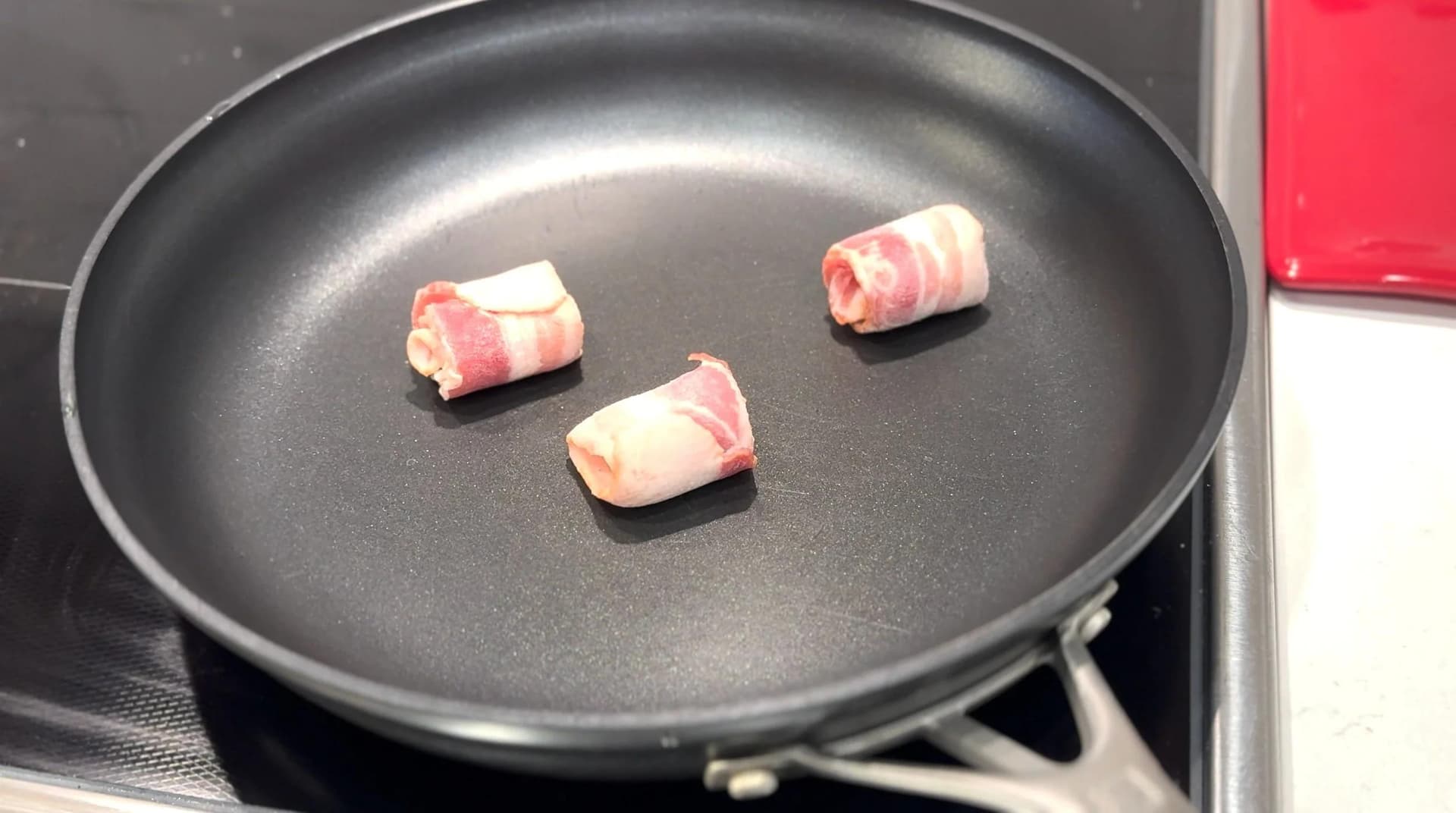 Three bacon strips cooking in a black skillet with water, showing the initial rendering stage on medium-low heat.
