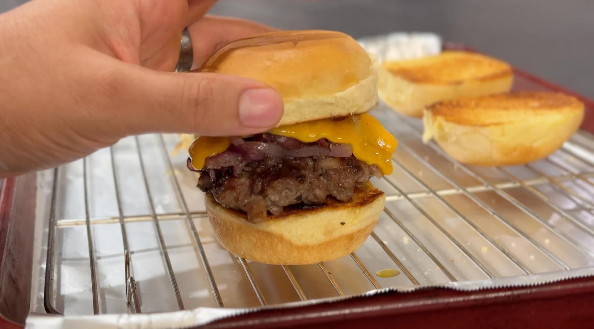 Hand holding a bacon cheeseburger slider with melted cheese and caramelized onions on a toasted bun.