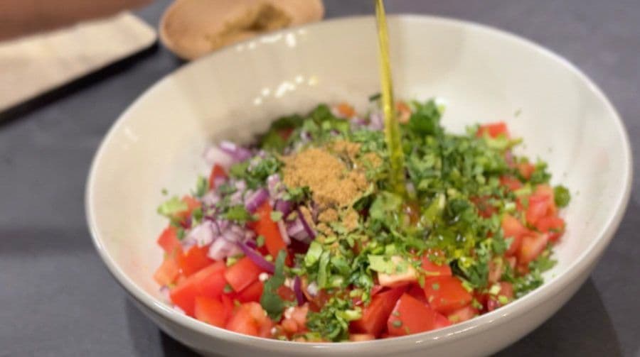 Olive oil being drizzled over fresh pico de gallo in a white bowl with diced tomatoes, cilantro, onions, and spices.
