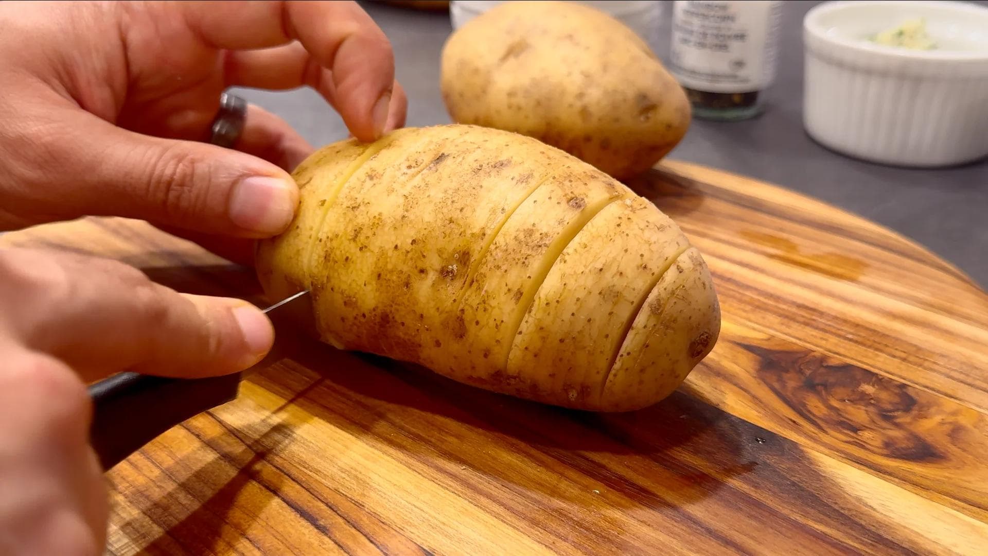 Hand slicing a potato three-quarters of the way through on a wooden cutting board for Hasselback potatoes.