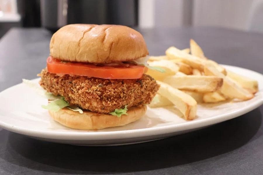 Crispy paneer tikki burger with tomato slices and lettuce on a bun, served with french fries on a white plate.