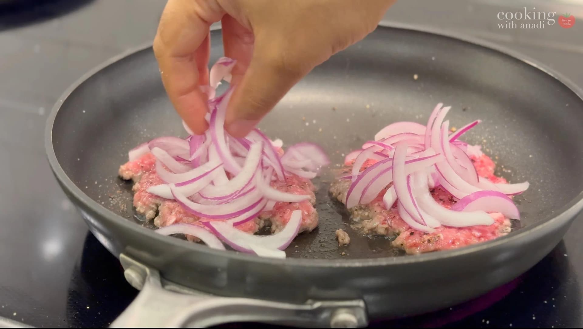 Hand adding sliced red onions to ground beef patties cooking in a skillet.