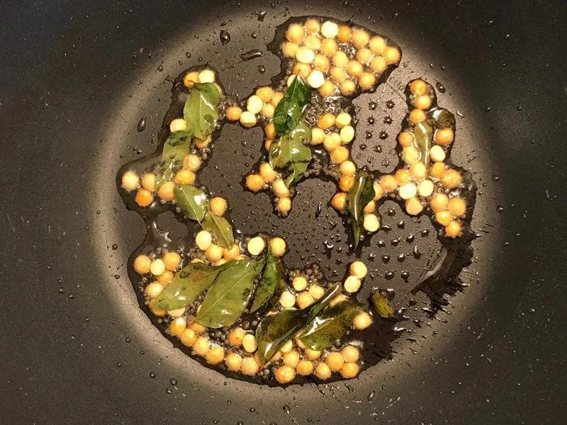Chana dal and curry leaves tempering in oil with mustard seeds in a dark pan, showing the initial seasoning step.