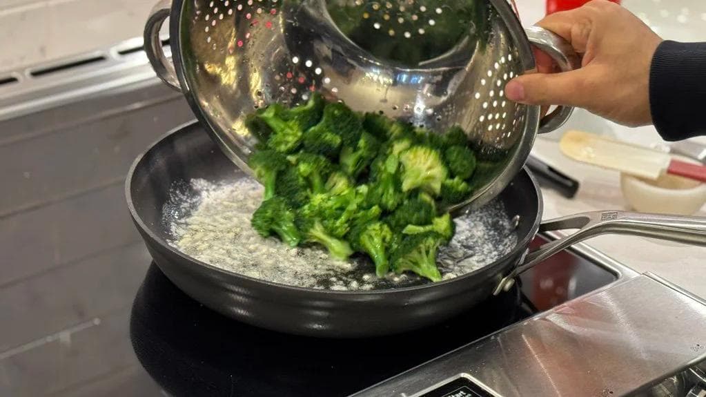 Straining cooked broccoli florets from a colander into a pan with garlic butter sauce on the stovetop.
