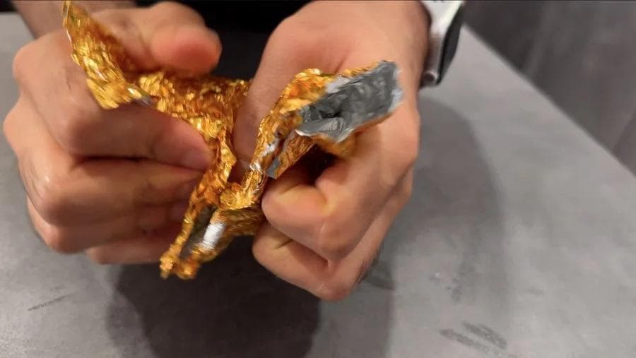 Hands holding a baked potato wrapped in gold foil being tested with a fork to show it's fully cooked inside.