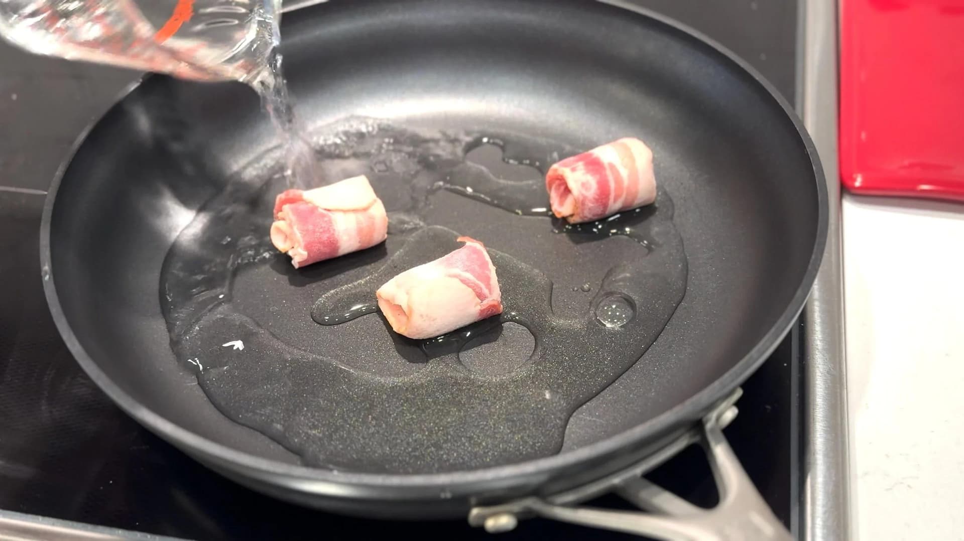 Bacon strips cooking in a black skillet with water being poured in, on a stovetop.