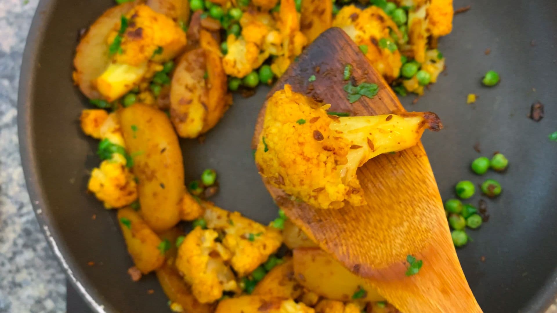Golden roasted potatoes and cauliflower with green peas in a dark skillet, wooden spoon stirring the curry.