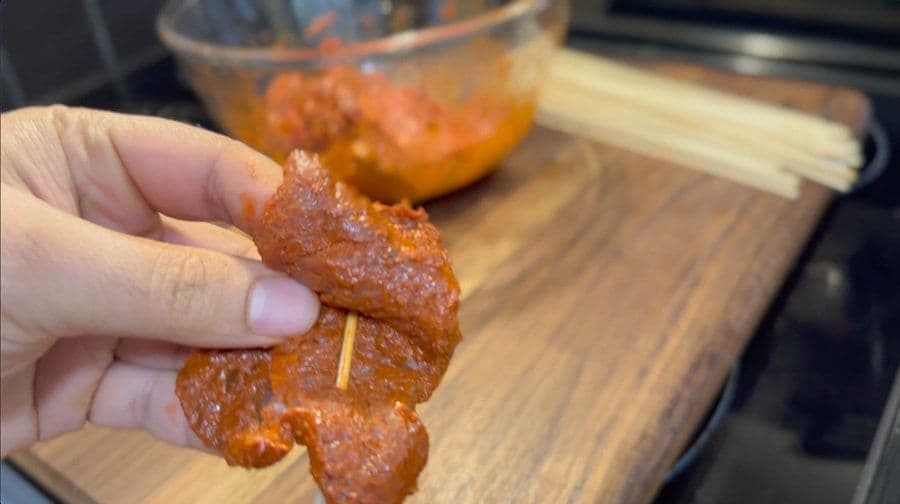 Hand holding a cooked Brazilian churrasco skewer with caramelized meat and a grill pan in the background.