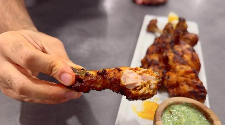 Hand holding a golden-brown tandoori chicken drumstick with a small bowl of green sauce on the side.