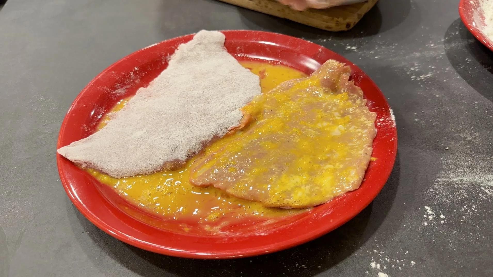 Breaded veal cutlet being dredged in flour on a red plate during schnitzel preparation.