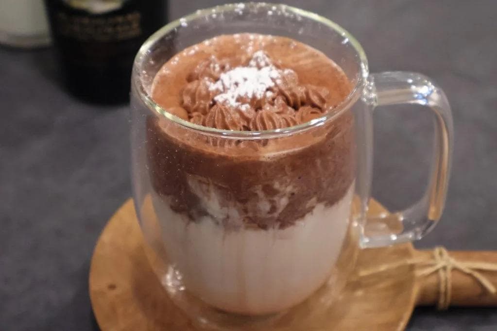 Rich whipped Baileys hot chocolate in a glass mug with layered chocolate and milk, topped with whipped cream and cocoa powder.
