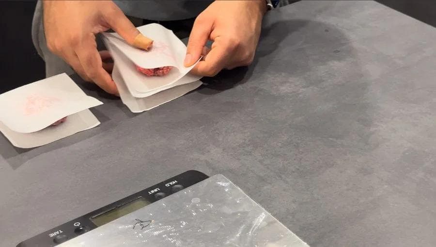 Hands placing a portion of ground beef onto a parchment paper square next to a kitchen scale.