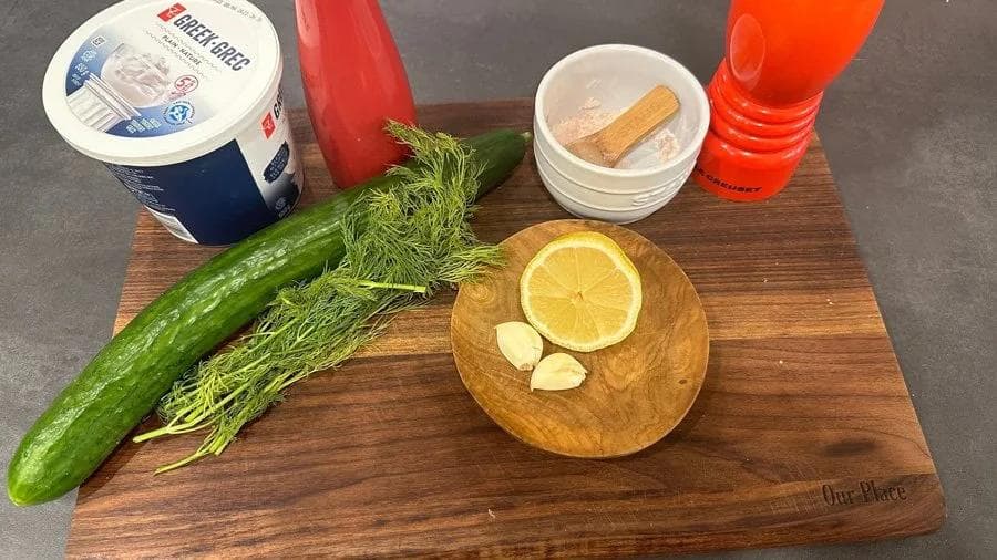 Ingredients for tzatziki sauce arranged on a wooden board: cucumber, fresh dill, lemon, garlic, Greek yogurt, olive oil, and spices.