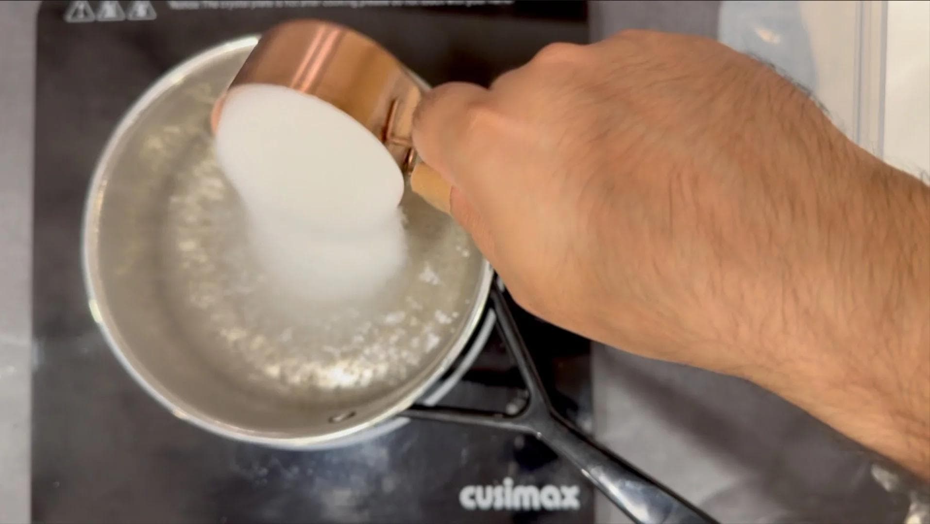 Pouring vanilla syrup from a copper measuring cup into a saucepan of dissolved sugar and water.