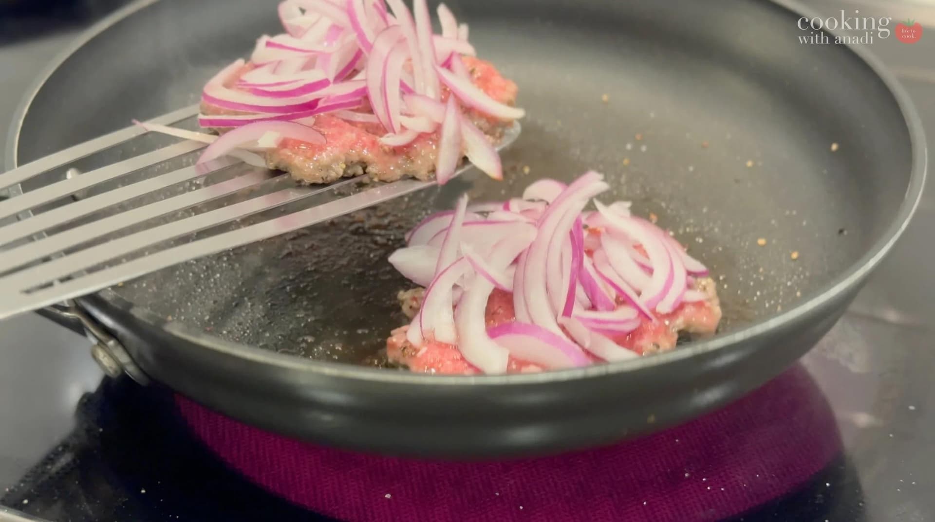 Two smash burger patties topped with sliced red onions cooking in a skillet with a spatula.