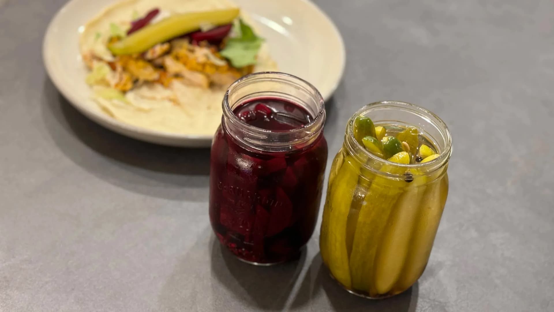 Two jars of homemade pickled vegetables with a plated dish in background featuring sliced vegetables on cream sauce.